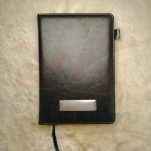 Black leatherette diary with a brushed silver metal plate for personalization on the cover, lying on a white, plush background